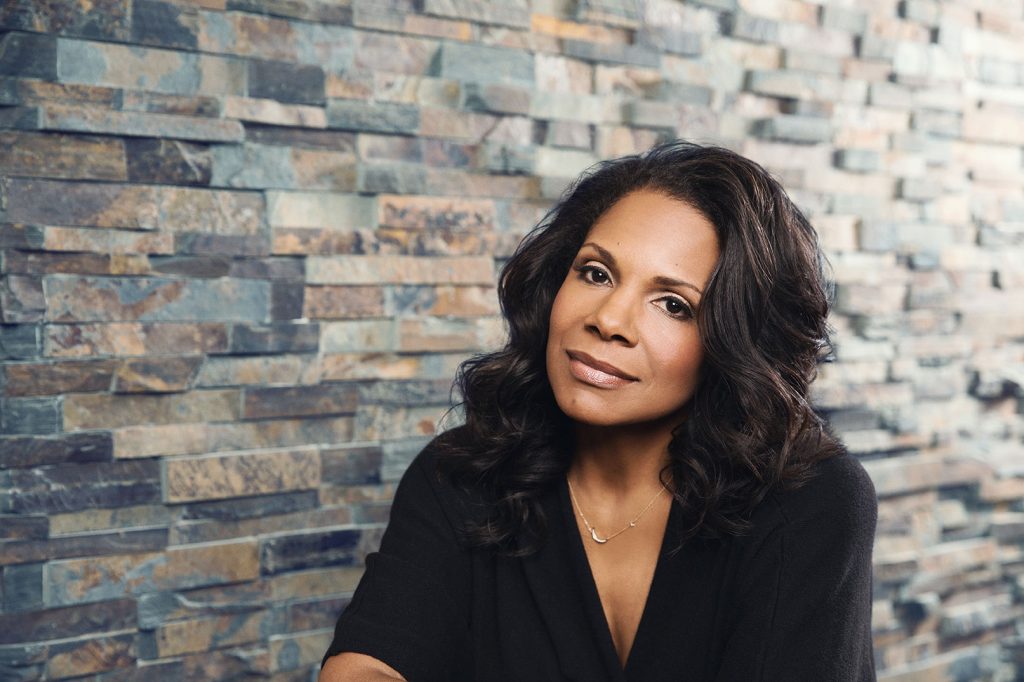 Audra McDonald sits in front of a multi-colored stone wall, wearing a black top and faintly smiling while looking toward the camera.