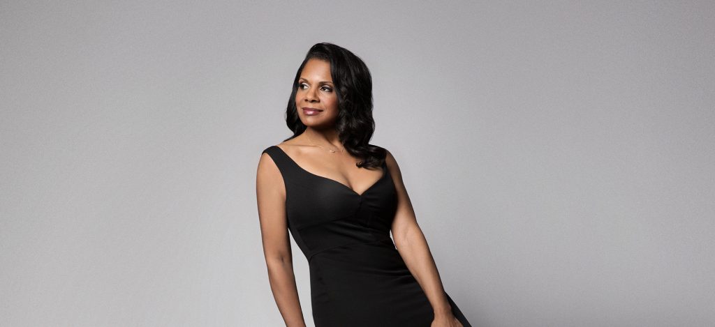 Audra McDonald sits on a stool wearing a form fitting black stress and looking off to the side.