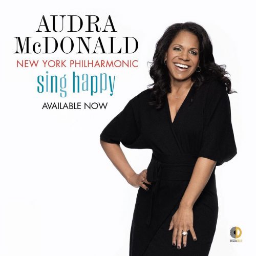 The album cover of "Sing Happy with the New York Philharmonic" featuring Audra McDonald wears a long black blouse with jeans, resting one hand on her hip and smiling.