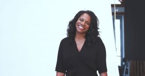 Audra McDonald smiles with her head tilted to the side, posing in front of a white background.