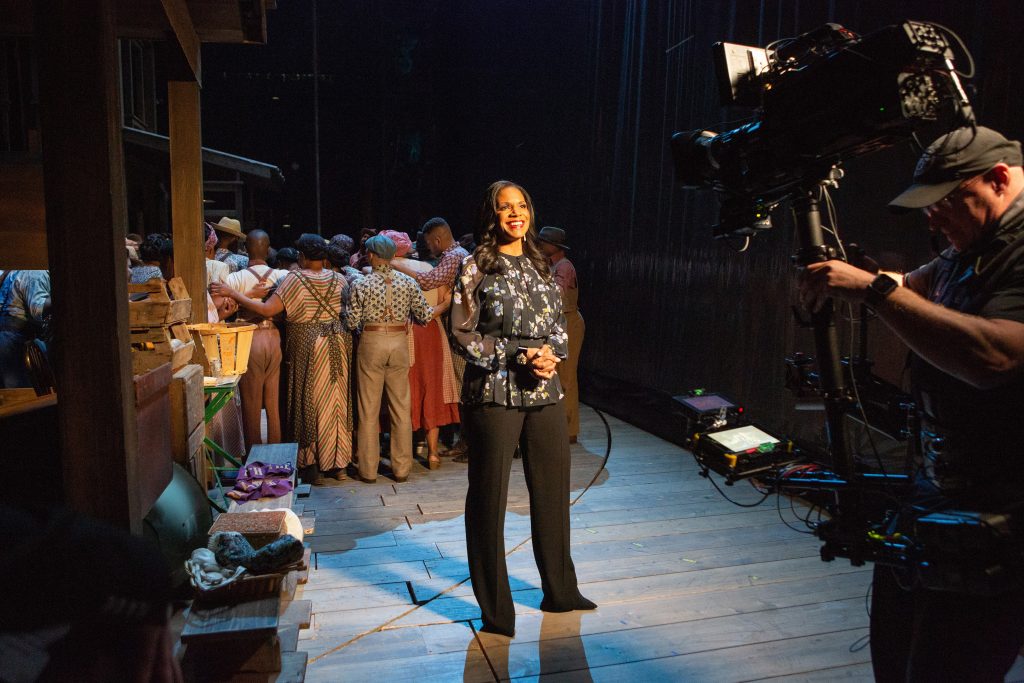 Audra McDonald hosts the Met Opera Live in HD of Porgy and Bess. She stands in front of a camera smiling in front the the cast who are about to go onstage.